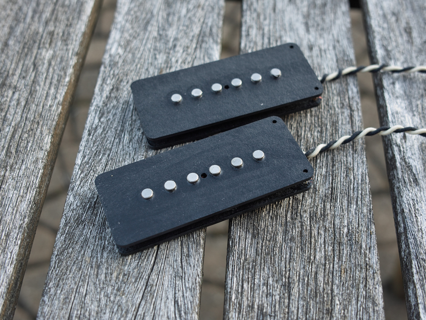Two Lake Surfer model guitar pickups for Jazzmaster style guitars. The pickups are placed on a wood table. They have black bobbins.